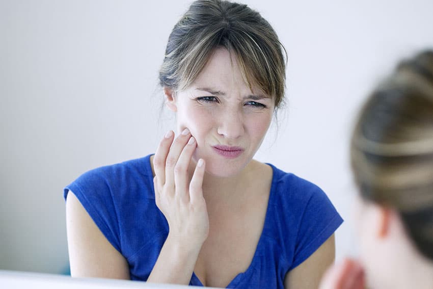 young woman standing in front of mirror, holding jaw because of TMJ pain